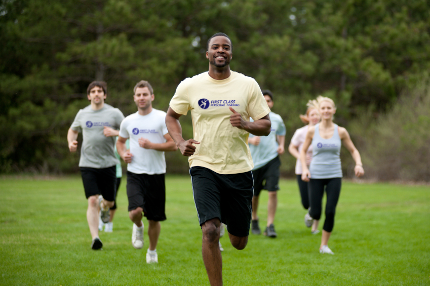 A man leads a group of people running through a field, they are all wearing matching white t-shirts with the First Class Personal Training logo on it.