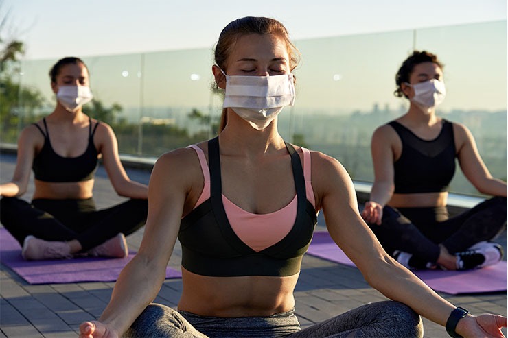 Three women sit cross legged on a balcony in a mediation pose. They are all wearing medical face masks.