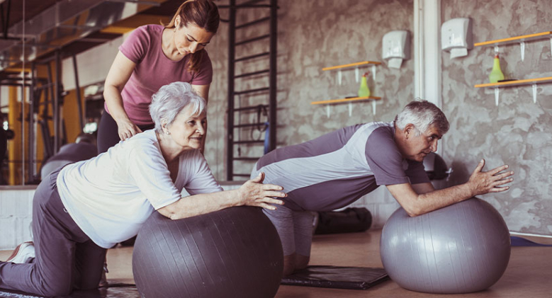 A personal trainer assists two elderly clients who are leaning on two exercise balls in a gym.