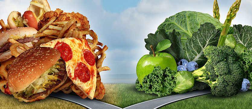 Two roads splitting off. The left side of the roads leads to pizza, burgers, french fries, and chicken. The right side leads to a pile of fresh green vegetables and blueberries.