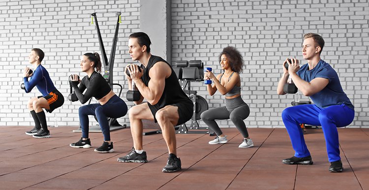 A group of 5 people in a gym doing squats holding dumbbells.
