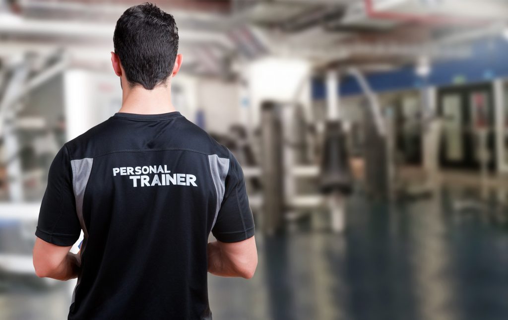 A man is in a gym standing with his back to the camera. He is wearing a black and white t-shirt with the words "Personal Trainer" on the back.