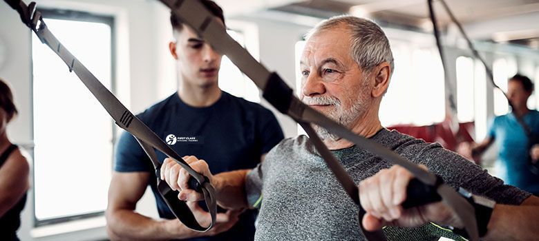 A personal trainer stand behind an elderly male patient. The trainer is assisting him with arm exercises.