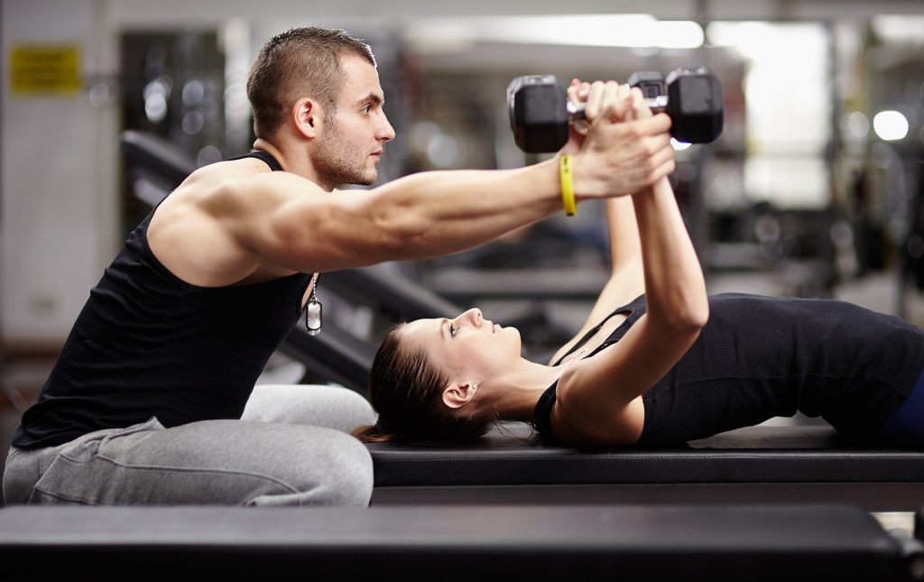 A personal trainer is sitting behind a woman laying on an exercise bench in a gym. She is lifting two metal dumbbells, supposed by the personal trainer.