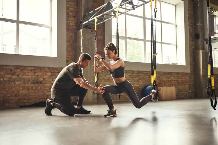 A male personal trainer coaches a female client doing a leg exercise. He is assisting the client doing lunges.