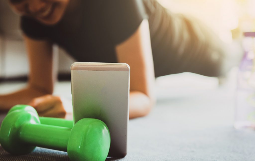 A phone stands up leaning against a green dumbbell. There is a person planking in the background smiling.