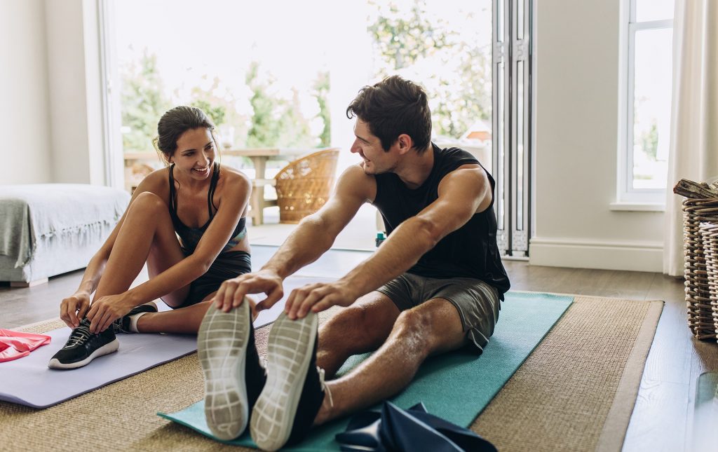 A woman sitting on a yoga mat ties her shoe as she smiles at a man beside her. The man is also sitting on a yoga mat and is stretching touching his toes.
