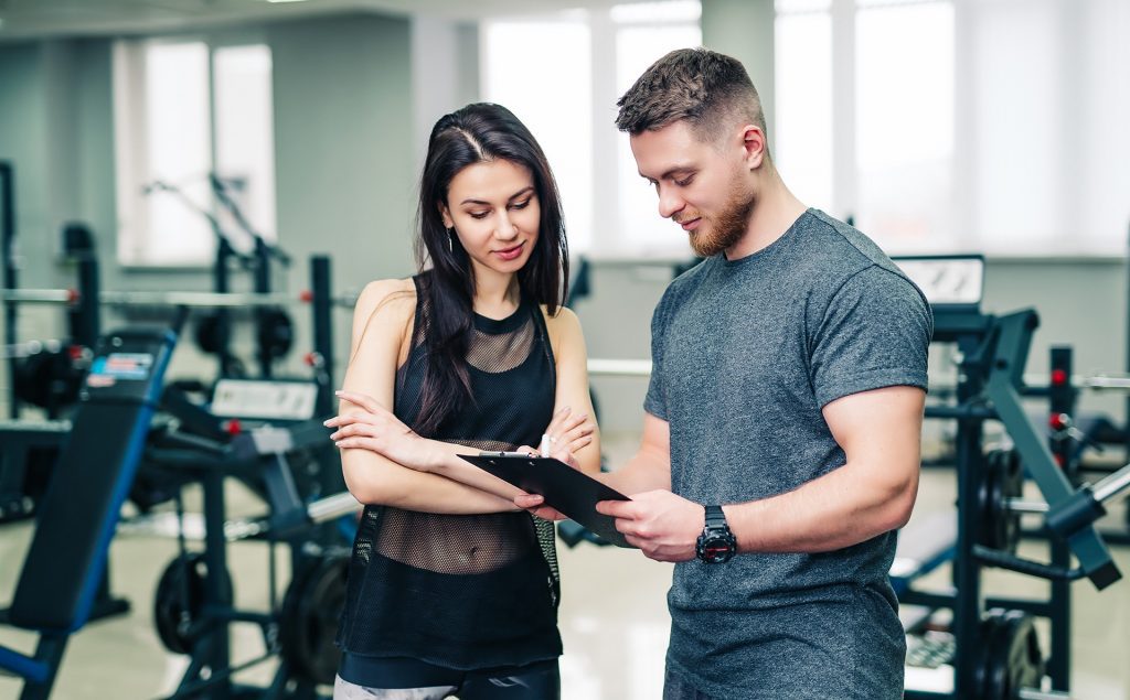 A man holds a clipboard and talks to a young woman, who is wearing gym clothes. They are standing together in a gym.