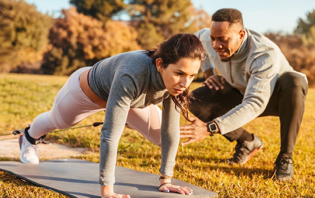 A woman has an elastic cord attached to her ankles. she is on her hands planking in the grass on a yoga mat. She is focused on exercising as her personal trainer rests his hands in front of her left knee, which is bent towards her chest.