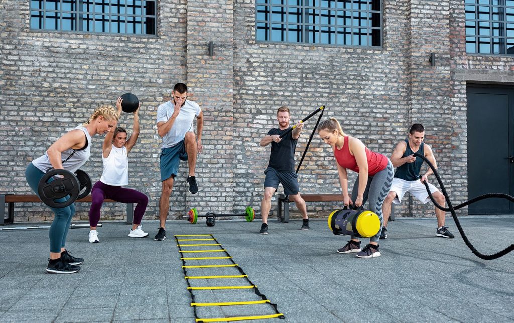 Six people in gym clothes are training outside. They are all using various gym equipment such as barbells, ropes, exercise balls, and floor ladders.