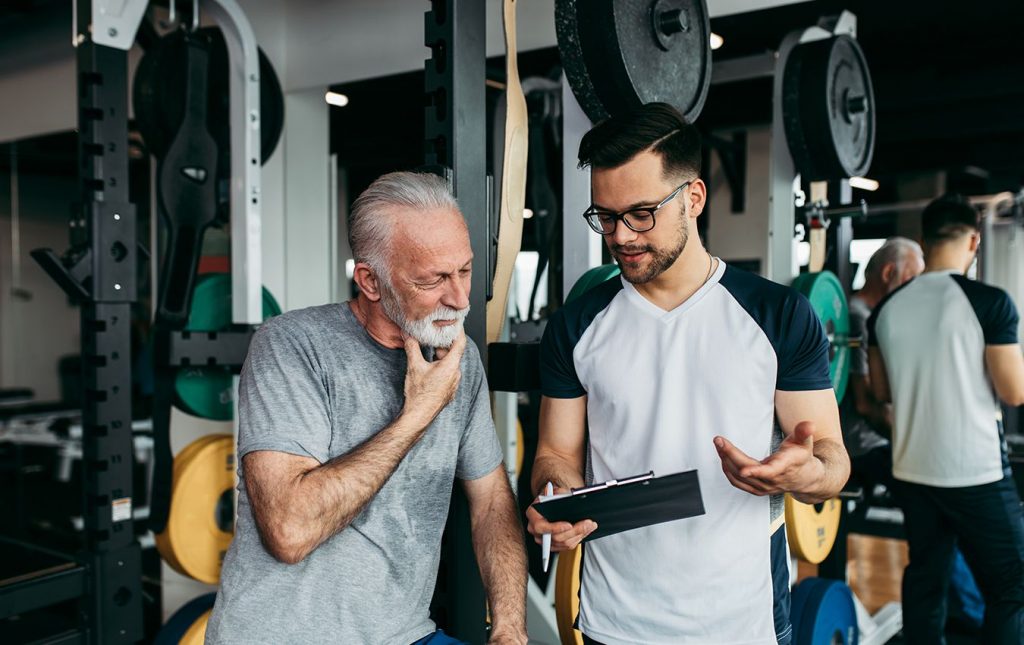 An elderly male with a beard and a personal trainer stand in a gym. The elderly male has his hand on his chin and is looking at a clipboard held by the personal trainer. The personal trainer is talking to the man.