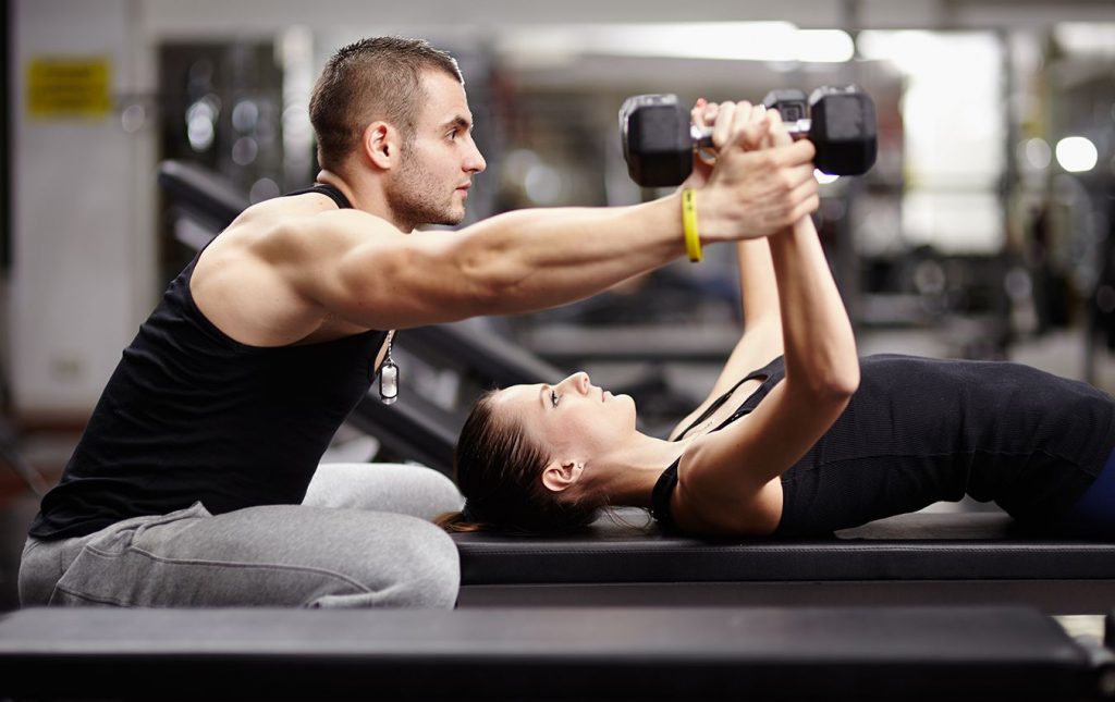 A woman lays on an exercise bench. She is on her back with her arms up holding two dumbbells. There is a personal trainer behind her. He is seated near her head and is holding her hands, supporting her holding the dumbbells.