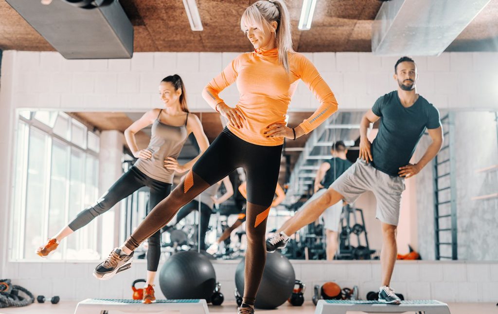 A woman stands in front of two people in a gym. All of the people have their hands on their hips with their leg extended sideways.