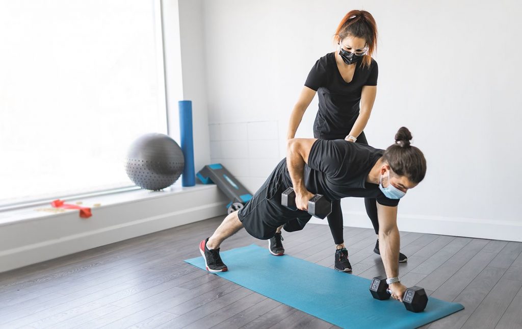 A man and woman are both wearing medical face masks. The woman is wearing safety glasses. The woman stands above the man assisting him in arm raises with two large dumbbells. He is on the floor in a plank on a yoga mat.