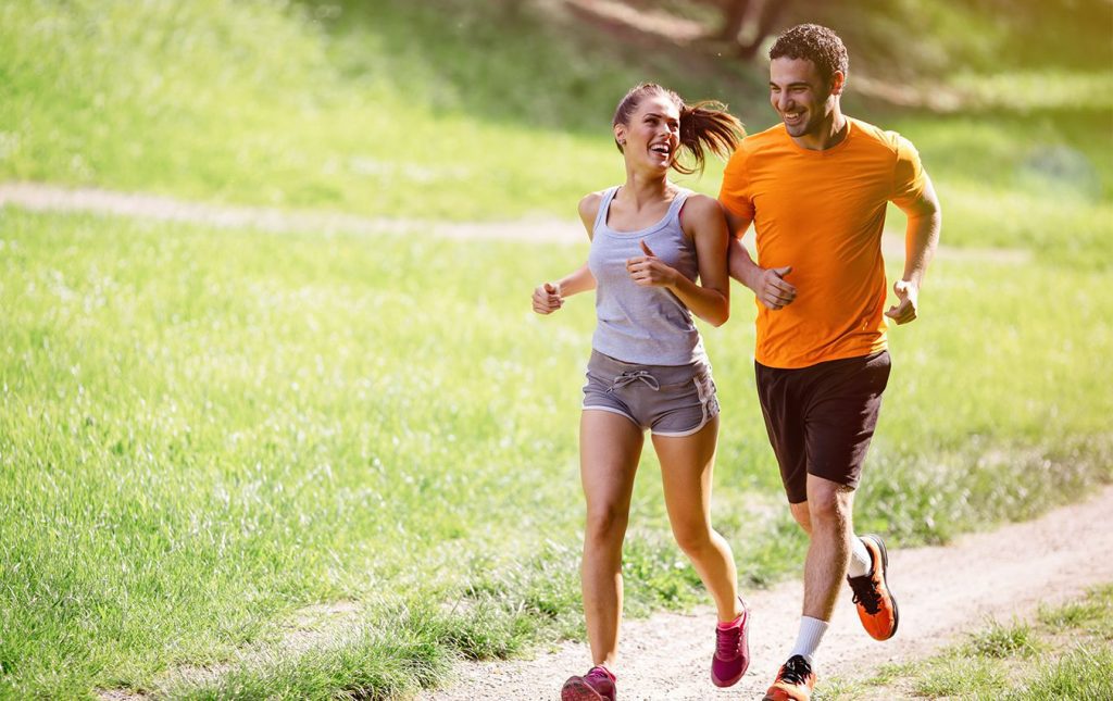 A man and woman smile at each other while running outdoors on a dirt trail, surrounded by grass.