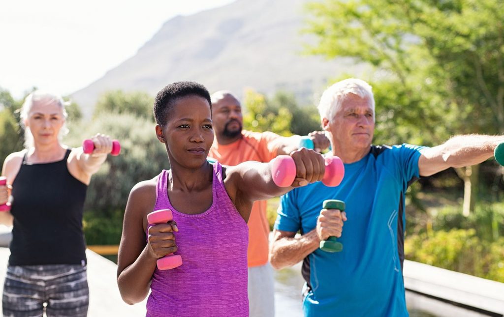 Four older people stand outside exercising. They are holding small colourful dumbbells with their lefts arms extended out holding the dumbbell.