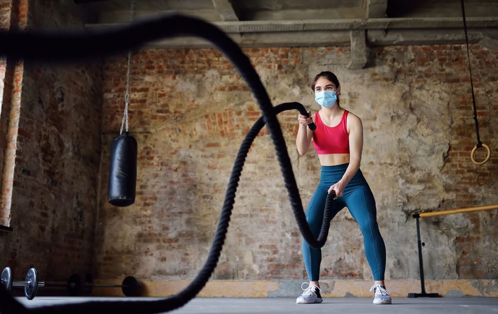 A young woman wearing a medical face masks lifting exercise ropes in a gym. There is exercise equipment behind her.
