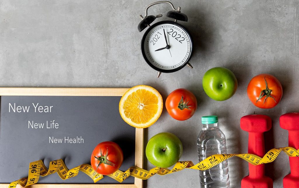 A chalkboard on the floor reads "New Year, New Life, New Health." A measuring tape, dumbbells, and a bottle of water lay beside the clipboard. A clock with the years 2021 and 2022 sit above a line of Apples, Oranges, and Tomatoes.. The arm on the clock is nearing 2022, signaling the start of a New Year.