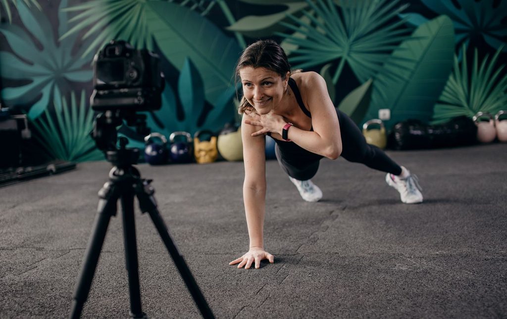 A woman planks on the ground in front of a camera on a tripod. Her left arm is bending up with her left hand touching her right shoulder. She is smiling at the camera.