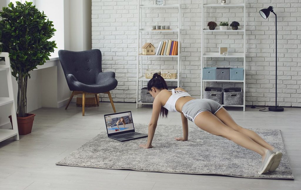 A woman wearing gym clothes exercises in her living room on a rug. She is planking in front of a laptop that shows an image of another woman planking.