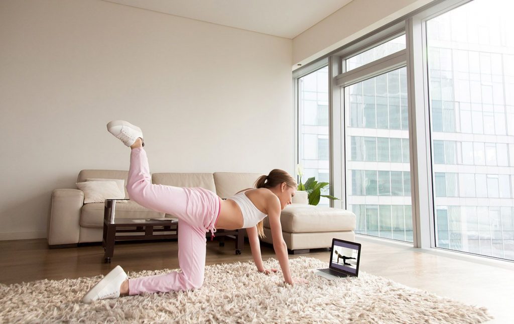 A woman exercising at home on a shag rug. She is watching an exercise video about doing a plank. She is on her hands with her right leg extended up in the air.