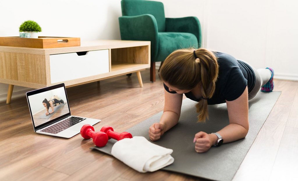 A woman planks on a yoga mat while watching a laptop. On the laptop, there is an image of a woman also in a plank. A towel lays on the floor in front of the woman, as well as two small red dumbbells.