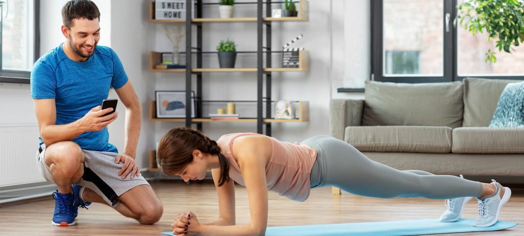 A woman planks on a living room floor on a yoga mat. A personal trainer is kneeling next to her smiling looking at a cellphone in his left hand.