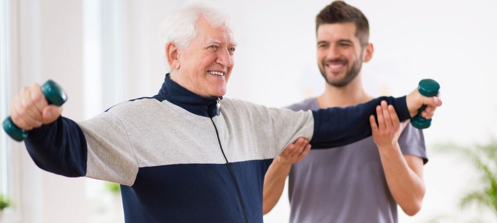 An elderly man smiles while lifting two small dumbbells out from his sides. A personal trainer smiles and helps the man lift his left arm with both hands.