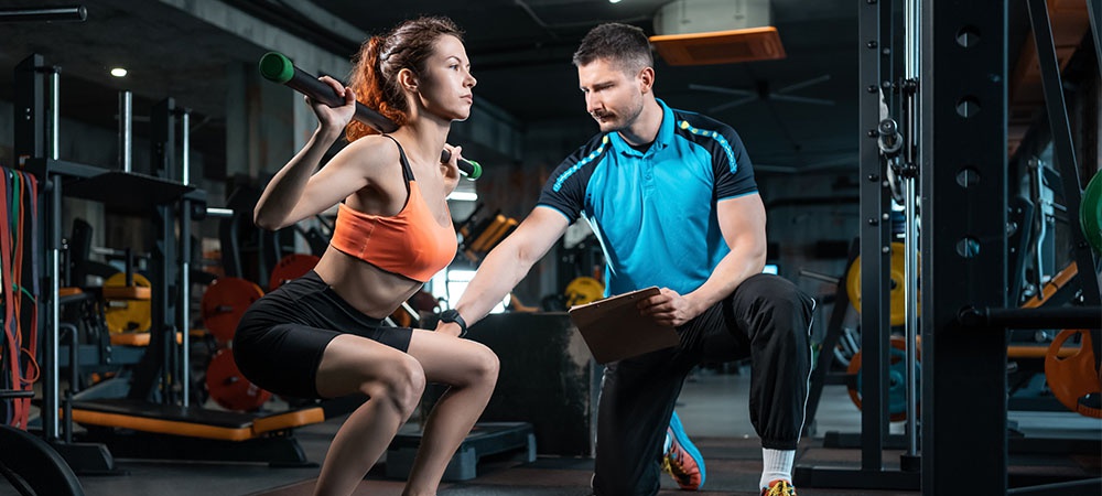 A woman in a gym is squatting with a long metal bar on her shoulders. A personal trainer kneels next to her holding a clipboard.
