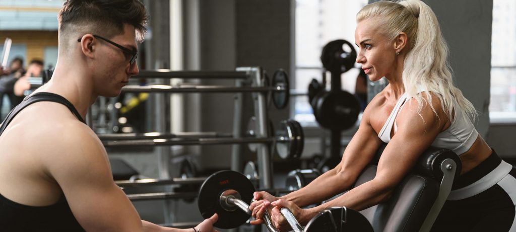 A woman leans her arms over a exercise bench in a gym. She is holding a large barbell and facing a man with glasses. The man is helping her lift the barbell