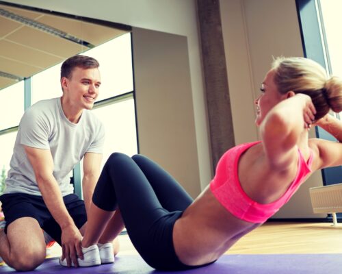 Doing Sit-Ups Is A Simple Bodyweight Exercise A young woman in workout attire does sit-ups with the guidance of a personal trainer.
